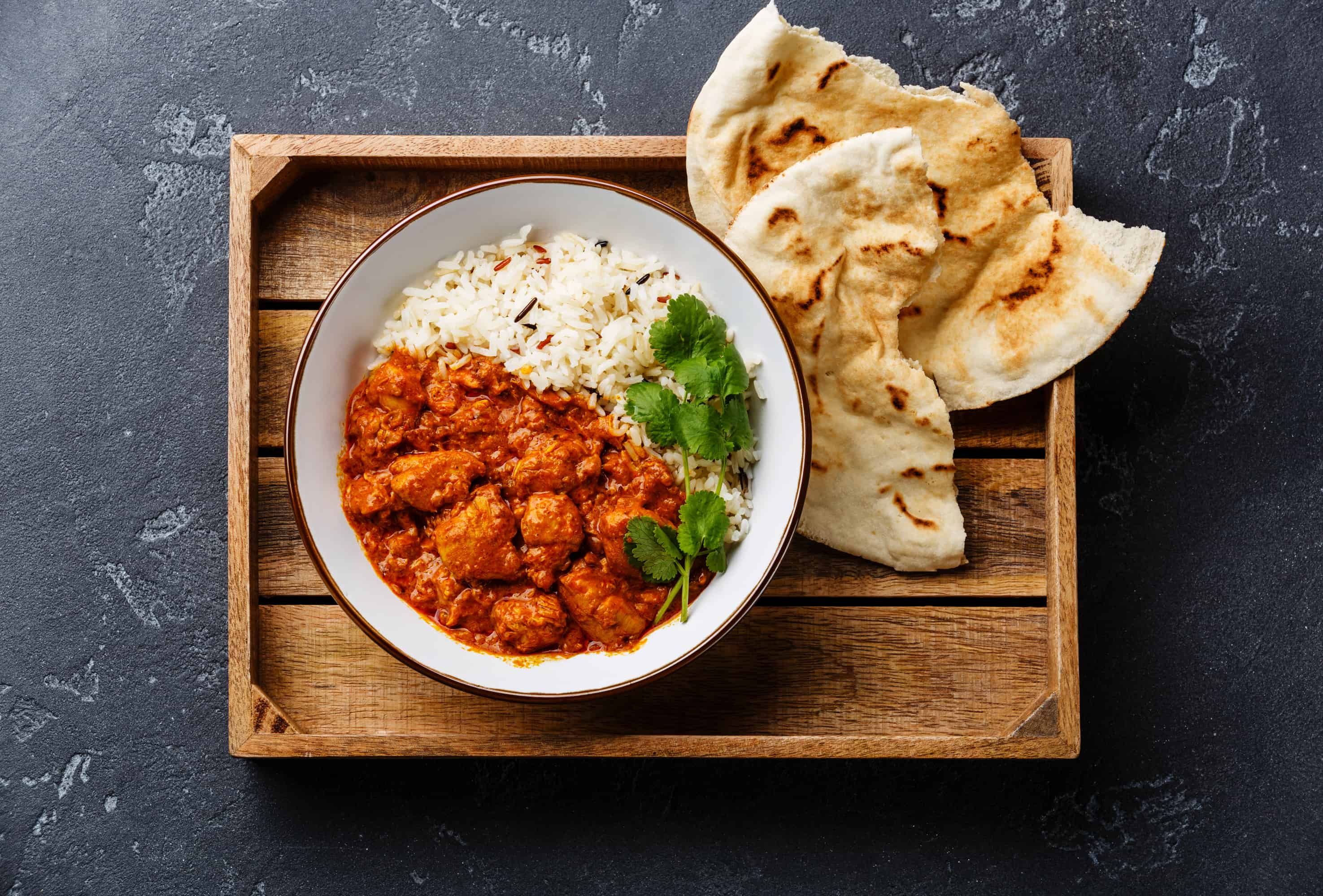bowl of rice and chicken tikka masala on wooden tray with naan bread