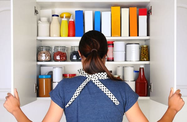 A woman seen from behind opening the doors to a fully stocked pantry. The cupboard is filled with various food stuff and groceries all with blank labels. Horizontal format the woman is unrecognizable.